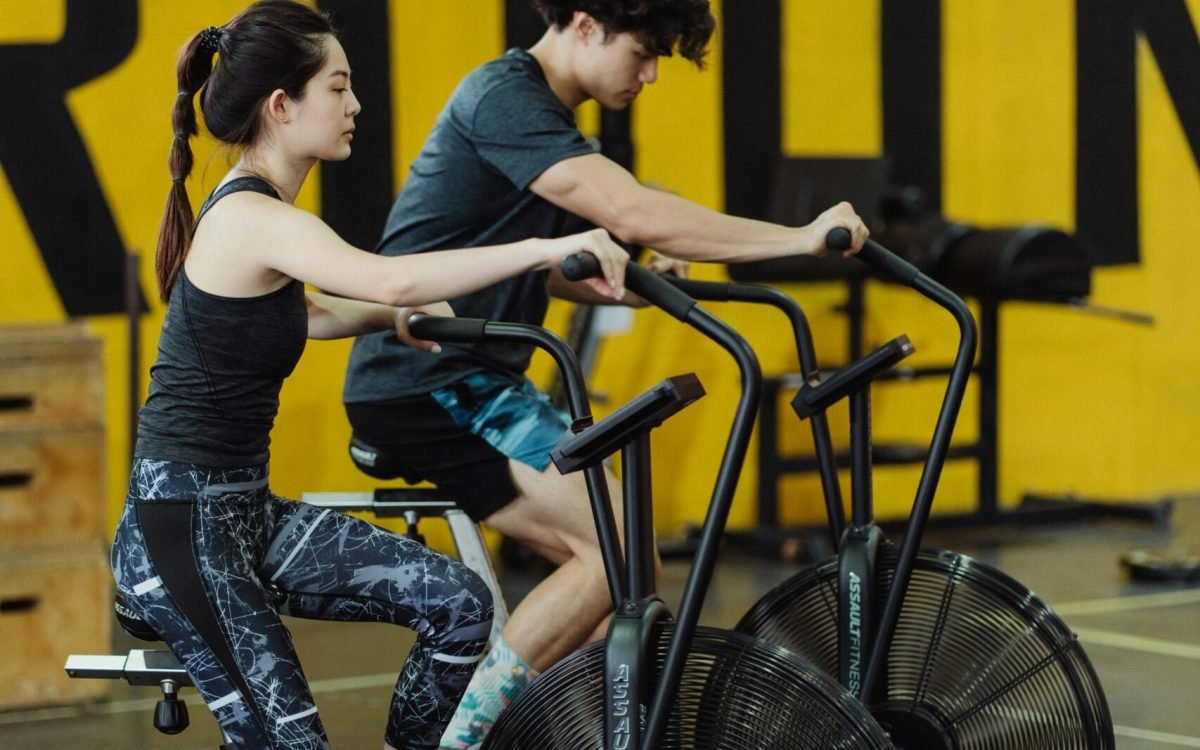 Asian couple in activewear working out on stationary bikes indoors, promoting fitness.